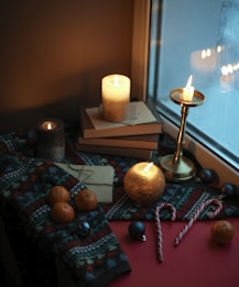 A cozy winter setting near a window, featuring lit candles on top of stacked books. A patterned knit blanket covers part of the scene, with several small oranges placed on it. Nearby are letters wrapped with string and candy canes. The warm glow from the candles is reflected in the window, enhancing the intimate ambiance.