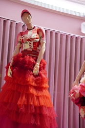 A mannequin is dressed in a vibrant outfit featuring a layered red and orange tulle skirt topped with a graphic-patterned top. The mannequin also wears a red cap and matching gloves. The background consists of soft, purple curtains, adding to the elegant display.