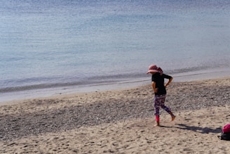 A child wearing a wide-brimmed pink hat with red polka dots, a black shirt, and colorful leggings walks barefoot on a sandy beach. The beach has pebbles scattered across it, and the calm sea stretches out to the horizon with gentle waves lapping at the shore. A small red and black bag is placed on the sand nearby.
