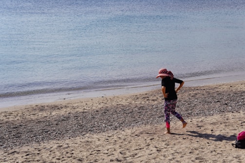 A child wearing a wide-brimmed pink hat with red polka dots, a black shirt, and colorful leggings walks barefoot on a sandy beach. The beach has pebbles scattered across it, and the calm sea stretches out to the horizon with gentle waves lapping at the shore. A small red and black bag is placed on the sand nearby.
