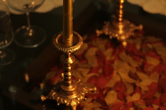 A brass candle holder stands elegantly on a table surrounded by red and yellow autumn leaves. The candle holder features intricate patterns and is paired with glassware in the background, suggesting a cozy and inviting atmosphere.