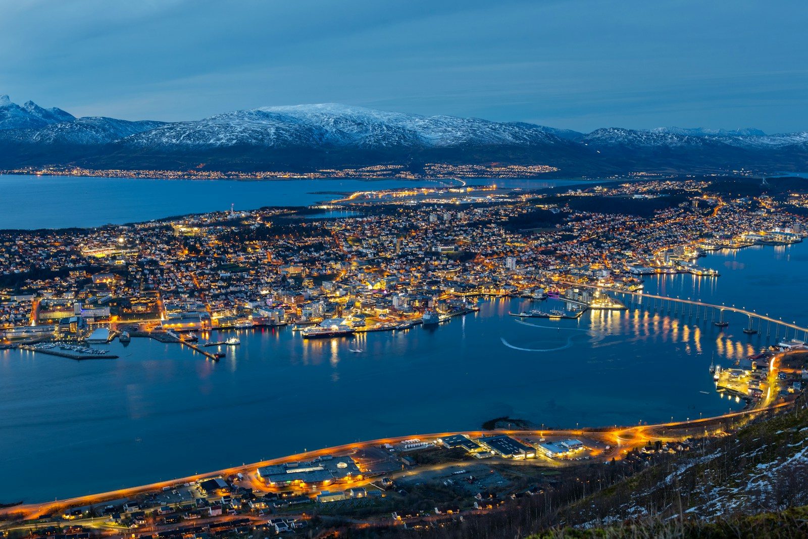 An aerial view of Tromsø at dusk, the city lights, the bridge over the sound and snow-covered ridges behind