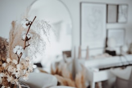 A soft-focused interior scene featuring dried flowers and cotton stems in the foreground. The background has blurred artwork on the walls and subtle, neutral-colored decor. The overall aesthetic is minimalistic and calming.