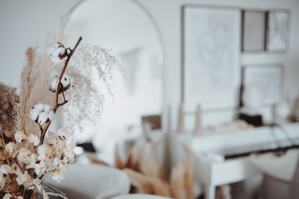 A soft-focused interior scene featuring dried flowers and cotton stems in the foreground. The background has blurred artwork on the walls and subtle, neutral-colored decor. The overall aesthetic is minimalistic and calming.