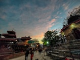 An evening or early morning scene at a cultural site with traditional architecture and pagoda-style roofs. People are casually walking around, and the sky features a blend of blue and orange hues, suggesting a sunset or sunrise.