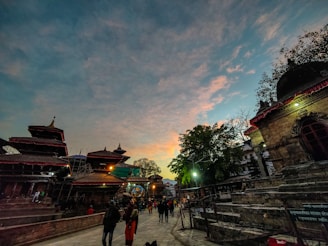 An evening or early morning scene at a cultural site with traditional architecture and pagoda-style roofs. People are casually walking around, and the sky features a blend of blue and orange hues, suggesting a sunset or sunrise.