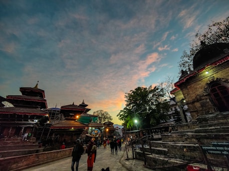 An evening or early morning scene at a cultural site with traditional architecture and pagoda-style roofs. People are casually walking around, and the sky features a blend of blue and orange hues, suggesting a sunset or sunrise.
