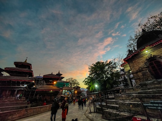 An evening or early morning scene at a cultural site with traditional architecture and pagoda-style roofs. People are casually walking around, and the sky features a blend of blue and orange hues, suggesting a sunset or sunrise.