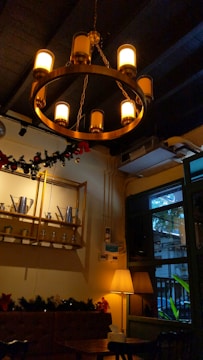 A warmly lit interior features a circular chandelier with multiple glowing bulbs. Below it, a wall shelf displays metal teapots and jars. A soft lamp casts a warm glow near a window overlooking greenery. Holiday garlands adorn the space, creating a cozy atmosphere.