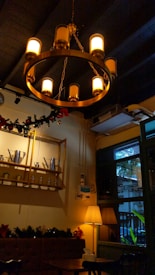 A warmly lit interior features a circular chandelier with multiple glowing bulbs. Below it, a wall shelf displays metal teapots and jars. A soft lamp casts a warm glow near a window overlooking greenery. Holiday garlands adorn the space, creating a cozy atmosphere.