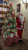 A beautifully decorated Christmas tree adorned with sparkling lights, red and gold ornaments, and red poinsettia flowers stands next to a life-sized Santa Claus figure holding a gift. A stack of wrapped presents lies at the base of the tree. The background features a wall with a framed picture and antlers, and a yellow shelf holding various items like cups and containers.