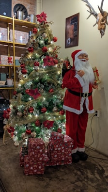 A beautifully decorated Christmas tree adorned with sparkling lights, red and gold ornaments, and red poinsettia flowers stands next to a life-sized Santa Claus figure holding a gift. A stack of wrapped presents lies at the base of the tree. The background features a wall with a framed picture and antlers, and a yellow shelf holding various items like cups and containers.