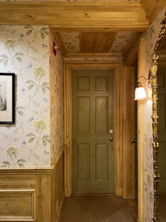 A hallway with a wooden door painted in muted green, surrounded by walls with floral wallpaper. The wallpaper features delicate patterns of flowers and butterflies in light hues. One wall has a framed picture, and there's an ornate gold-framed mirror on another. A vintage-style sconce light fixture casts a warm glow.