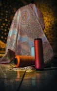 A richly patterned, multicolored fabric with intricate designs drapes over an object, set against a softly lit background. In front of the fabric, three large spools of yarn in orange, blue, and red hues are placed on the tiled floor, reflecting the ambient light.
