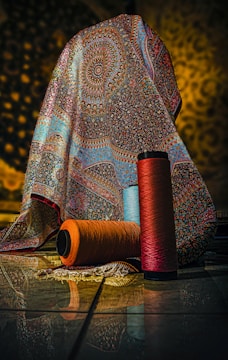 A richly patterned, multicolored fabric with intricate designs drapes over an object, set against a softly lit background. In front of the fabric, three large spools of yarn in orange, blue, and red hues are placed on the tiled floor, reflecting the ambient light.