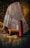 A richly patterned, multicolored fabric with intricate designs drapes over an object, set against a softly lit background. In front of the fabric, three large spools of yarn in orange, blue, and red hues are placed on the tiled floor, reflecting the ambient light.
