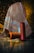 A richly patterned, multicolored fabric with intricate designs drapes over an object, set against a softly lit background. In front of the fabric, three large spools of yarn in orange, blue, and red hues are placed on the tiled floor, reflecting the ambient light.