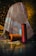 A richly patterned, multicolored fabric with intricate designs drapes over an object, set against a softly lit background. In front of the fabric, three large spools of yarn in orange, blue, and red hues are placed on the tiled floor, reflecting the ambient light.