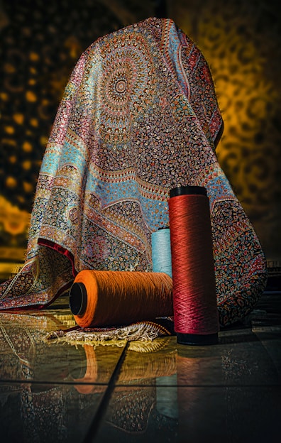 A richly patterned, multicolored fabric with intricate designs drapes over an object, set against a softly lit background. In front of the fabric, three large spools of yarn in orange, blue, and red hues are placed on the tiled floor, reflecting the ambient light.