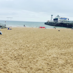 A sandy beach with a few people scattered across it, leading to a calm ocean in the background. A pier extends into the water, with a building labeled 'Restaurant' on it. The sky is overcast with clouds, and a couple of red signs are visible near the water's edge.