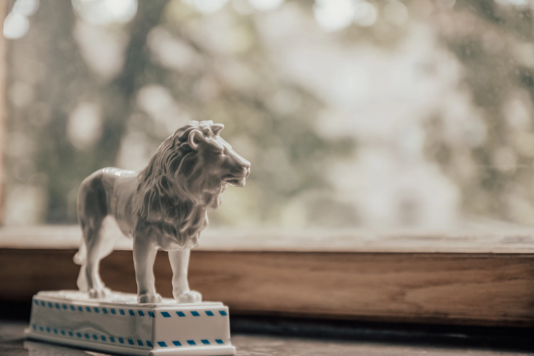 A white porcelain lion standing on a blue-banded plinth on a wooden windowsill