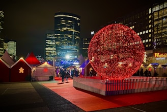 A festive outdoor scene featuring a large red illuminated spherical structure surrounded by a white picket fence. The area is populated with people walking under colorful lights and attending small hut-like structures with red roofs, decorated with glowing snowflake patterns. The background reveals tall skyscrapers with illuminated windows contrasting against the night sky.