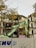 A partially demolished building with exposed rooms and floors is captured. A green excavator is in the foreground, actively engaged in tearing down the structure. The area is enclosed by a barrier that has graffiti on it. Tree branches with yellow leaves are visible at the top left corner.