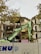 A partially demolished building with exposed rooms and floors is captured. A green excavator is in the foreground, actively engaged in tearing down the structure. The area is enclosed by a barrier that has graffiti on it. Tree branches with yellow leaves are visible at the top left corner.