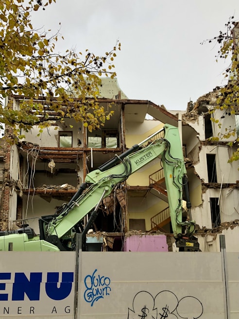 A partially demolished building with exposed rooms and floors is captured. A green excavator is in the foreground, actively engaged in tearing down the structure. The area is enclosed by a barrier that has graffiti on it. Tree branches with yellow leaves are visible at the top left corner.