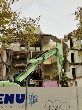 A partially demolished building with exposed rooms and floors is captured. A green excavator is in the foreground, actively engaged in tearing down the structure. The area is enclosed by a barrier that has graffiti on it. Tree branches with yellow leaves are visible at the top left corner.