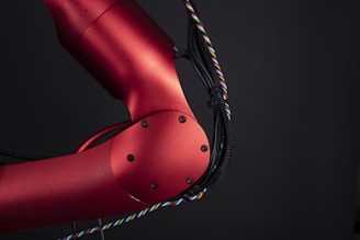 A close-up view of a red metallic robotic arm with visible joints and screws. The arm is connected with black and multicolored wires, set against a dark background, highlighting its mechanical and industrial design.