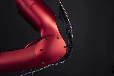 A close-up view of a red metallic robotic arm with visible joints and screws. The arm is connected with black and multicolored wires, set against a dark background, highlighting its mechanical and industrial design.