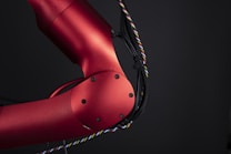 A close-up view of a red metallic robotic arm with visible joints and screws. The arm is connected with black and multicolored wires, set against a dark background, highlighting its mechanical and industrial design.
