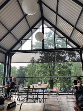 A spacious indoor cafe seating area with a high, triangular ceiling and large glass windows offering a view of lush green trees outside. People are seated at various modern wire-frame tables and chairs. There are minimalistic decorations and reserved signs on some tables.