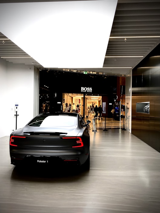 A sleek, modern car is parked indoors on a polished floor, with a glossy black and silver color scheme. The car's design is characterized by sharp lines and illuminated rear lights. In the distance, a clothing store with a prominent brand sign is visible, with people walking nearby.