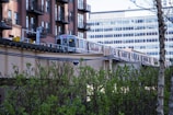 A train travels on an elevated track next to a row of modern brick apartment buildings with metal balconies. Behind the train is a large, multi-story office building with numerous windows. In the foreground, there are green bushes and a tree, contributing to an urban landscape with a hint of nature.
