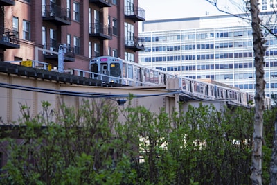 A train travels on an elevated track next to a row of modern brick apartment buildings with metal balconies. Behind the train is a large, multi-story office building with numerous windows. In the foreground, there are green bushes and a tree, contributing to an urban landscape with a hint of nature.
