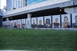 A mural featuring stylized portraits of diverse people painted on panels beneath a modern building. The mural displays a variety of individuals with different features and expressions, standing side by side. The backdrop includes a grassy area in the foreground and an urban architectural structure with reflective windows in the background.