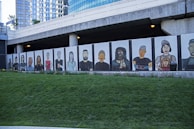 A mural featuring stylized portraits of diverse people painted on panels beneath a modern building. The mural displays a variety of individuals with different features and expressions, standing side by side. The backdrop includes a grassy area in the foreground and an urban architectural structure with reflective windows in the background.