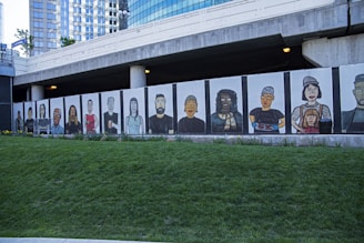A mural featuring stylized portraits of diverse people painted on panels beneath a modern building. The mural displays a variety of individuals with different features and expressions, standing side by side. The backdrop includes a grassy area in the foreground and an urban architectural structure with reflective windows in the background.