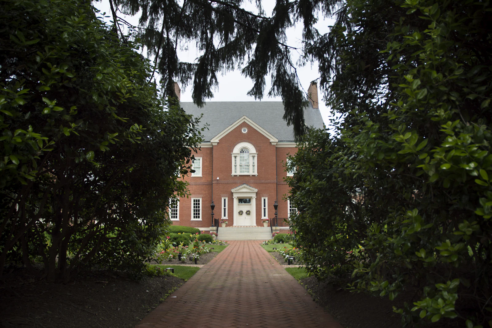 A red-brick colonial-style house at the end of a brick walkway, framed by tall trees
