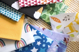 A collection of patterned fabric pieces in various designs including geometric shapes, polka dots, and animal motifs. The fabrics are arranged in an overlapping manner on a grid-patterned surface.