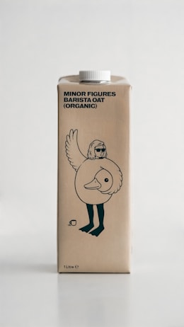 A carton of organic barista oat milk featuring a whimsical illustration of a bird with human-like legs wearing sunglasses. The packaging is simple with minimal text and design elements, set against a neutral background.