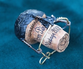 A wine or champagne cork engraved with the words 'Vintage 2002' and 'Champagne' held in a metal wire cage. The top of the cork is covered with black foil, and it rests against a teal fabric background.