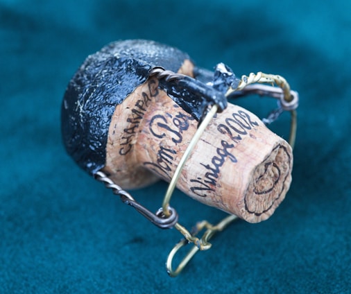 A wine or champagne cork engraved with the words 'Vintage 2002' and 'Champagne' held in a metal wire cage. The top of the cork is covered with black foil, and it rests against a teal fabric background.