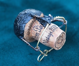 A wine or champagne cork engraved with the words 'Vintage 2002' and 'Champagne' held in a metal wire cage. The top of the cork is covered with black foil, and it rests against a teal fabric background.
