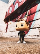 A Funko Pop figure stands on a rough-textured surface in front of a brick wall adorned with colorful mural art, featuring red, blue, and white geometric shapes.