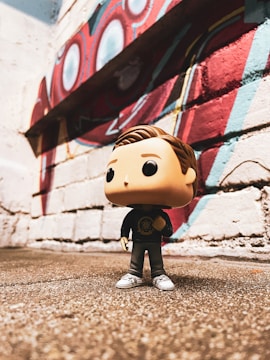 A Funko Pop figure stands on a rough-textured surface in front of a brick wall adorned with colorful mural art, featuring red, blue, and white geometric shapes.