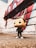 A Funko Pop figure stands on a rough-textured surface in front of a brick wall adorned with colorful mural art, featuring red, blue, and white geometric shapes.