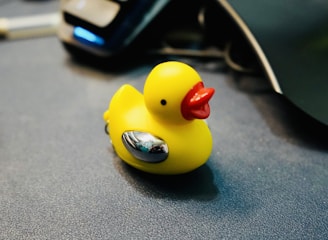 A yellow rubber duck with red beak and silver accents is placed on a dark surface with computer equipment visible in the background.